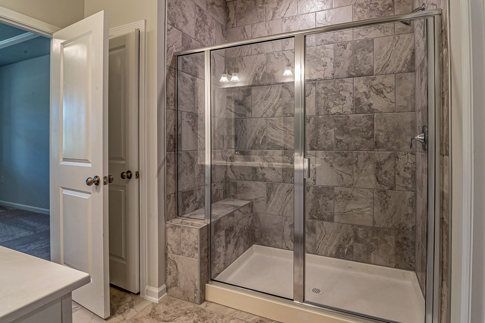 Shower with sliding glass doors, gray stone-pattern tiles, a built-in bench, and a white shower base.