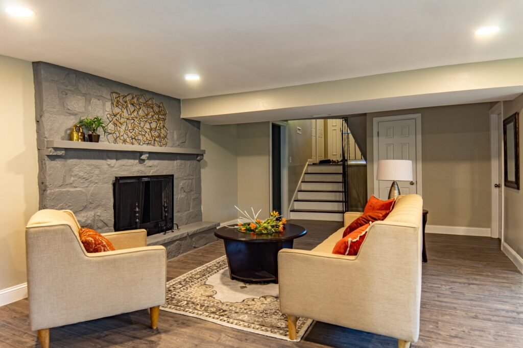 Lower-level living room with a gray stone fireplace, cream seating, orange cushions, and recessed lighting.