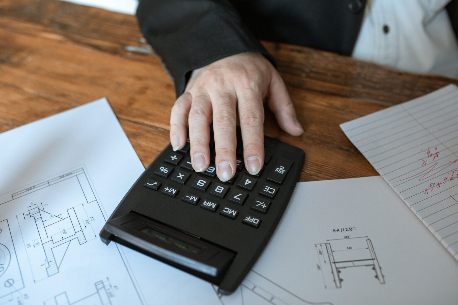 Person using a calculator beside technical drawings and handwritten notes on a wooden desk.